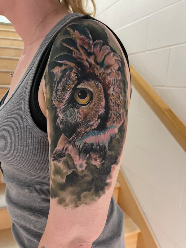 Color realism tattoo of a brown owl with green clouds on a woman's upper arm.