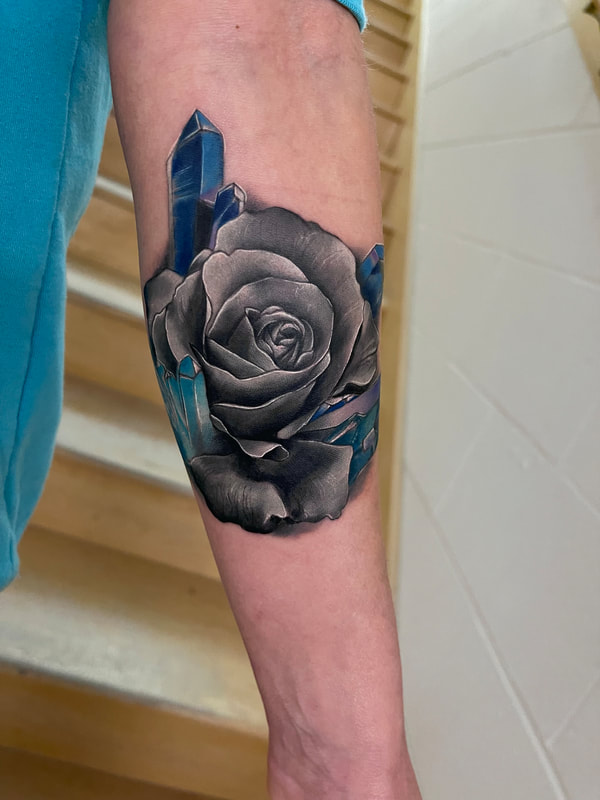 Black and grey color infused tattoo of a rose with purple crystals on a woman's inner arm.