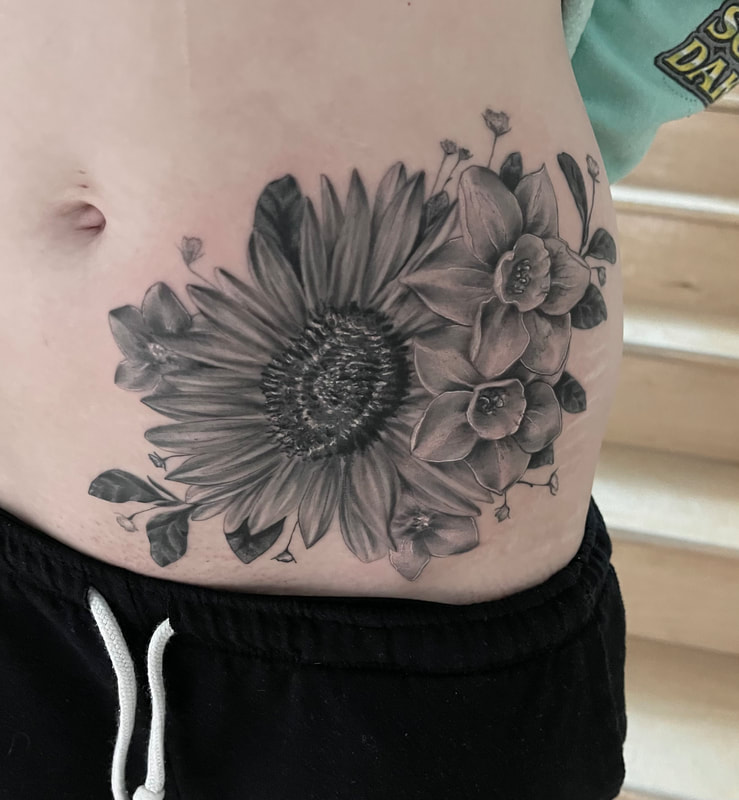 Black and grey realistic tattoo of a large sunflower and two daffodils on a woman's hip and stomach.