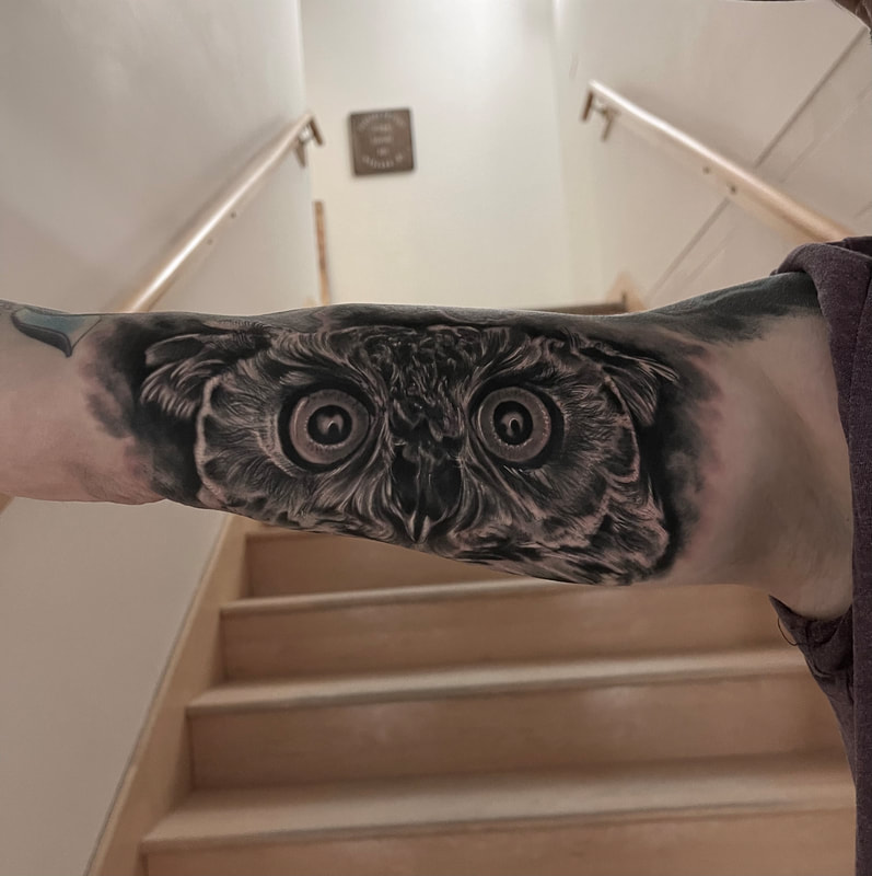 Black and grey realistic tattoo of an owl's face on an upper inner arm.