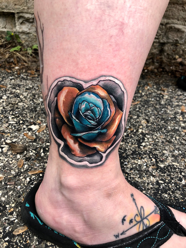 Sticker tattoo of a black and grey, copper, and blue rose on an ankle.