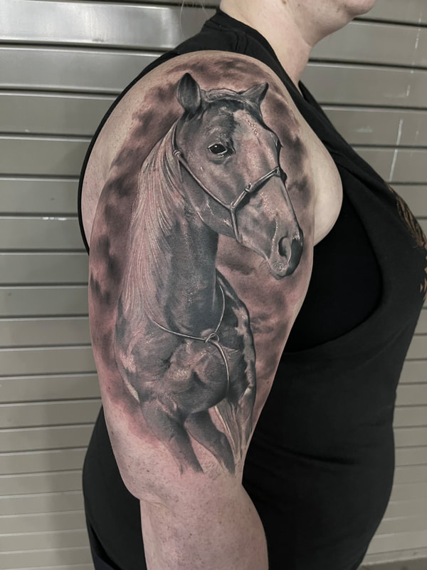 Black and grey realistic tattoo of a horse with a cloudy background on a woman's upper arm.