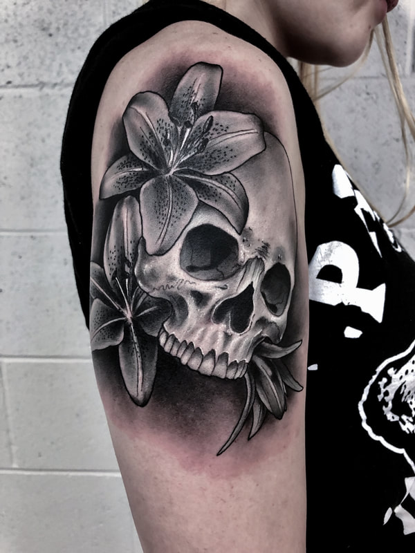 Black and grey realistic skull tattoo with lily flowers on an upper arm.
